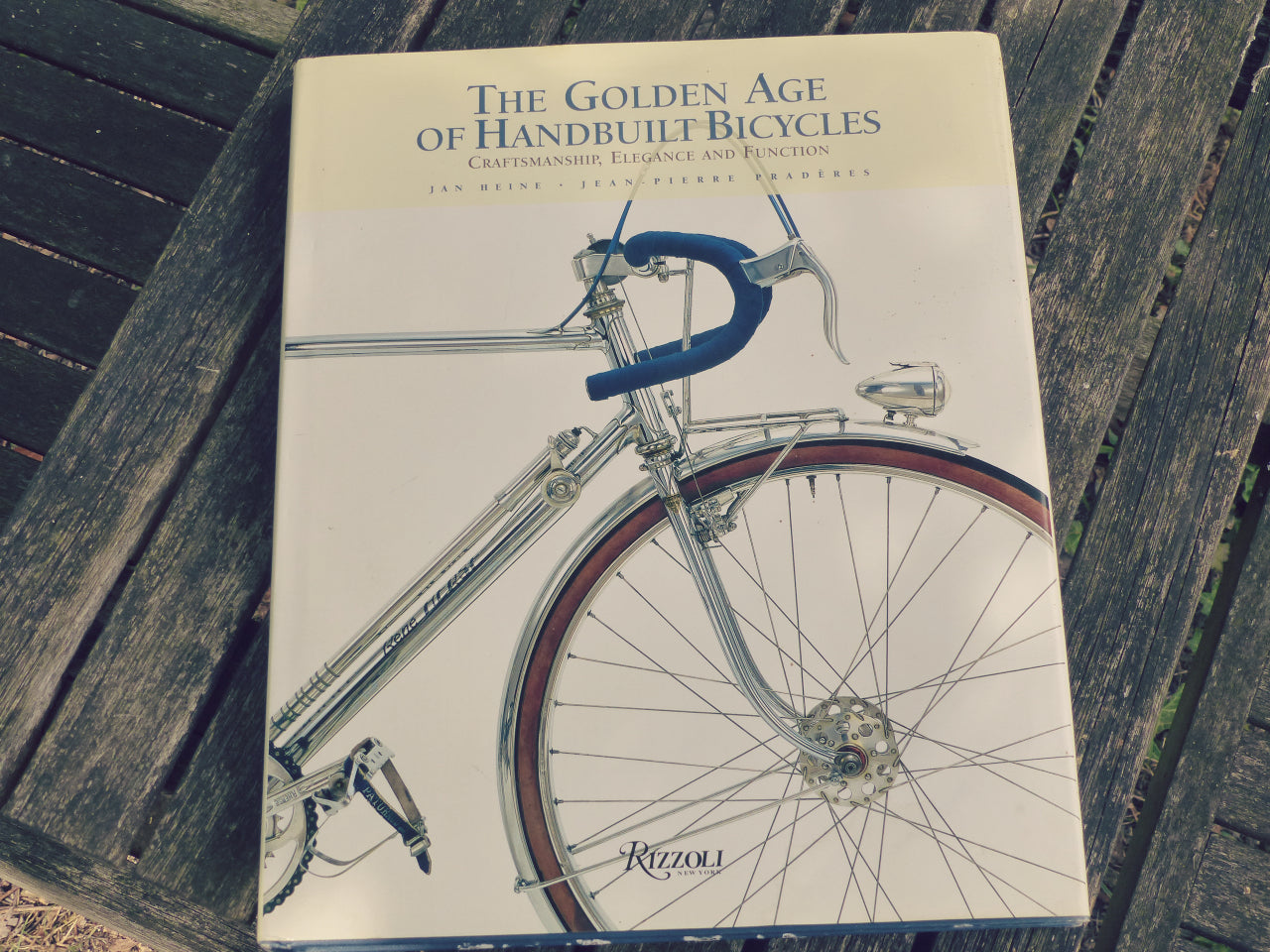 A book titled "The Golden Age of Handbuilt Bicycles" lying on a weathered wooden plank surface. The cover features a vintage bicycle illustration with a cream background, blue handlebars, and metallic silver frame details.